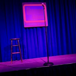 View of stage from 2nd row - The image shows a stage at The Comedy Studio in Harvard Square, with a wooden stool and a microphone on a stand positioned in front of a blue curtain. A sign with the venue's name is visible on the curtain, indicating the location as a comedy club. The stage is lit with purple and blue lighting, creating a vibrant atmosphere suitable for stand-up performances.
