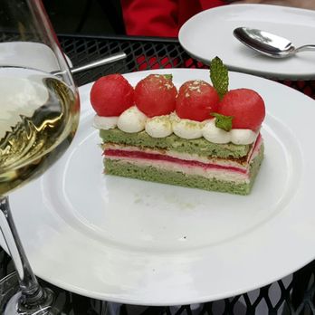 501 Local - Watermelon Cake - A slice of watermelon-themed cake with green sponge layers, pink filling, and white cream decorations is presented on a white plate. The cake is topped with watermelon balls, a small mint leaf, and dollops of cream. A glass of white wine and a spoon on a small white plate are visible nearby, set on a black metal table outdoors. - New American Near Me - Winnetka, Illinois
