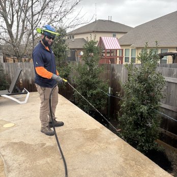 Moon Valley Nurseries - Angel, a man who takes what he does very seriously and makes his company look good! - A man wearing safety glasses, gloves, and a wide-brimmed hat waters plants in a backyard garden using a hose. He is standing on a concrete patio next to a garden bed with bushes and trees. The background features a wooden fence, neighboring houses, and a cloudy sky. The scene depicts outdoor gardening activity in a residential area. - Nurseries & Gardening Near Me - Buda, Texas