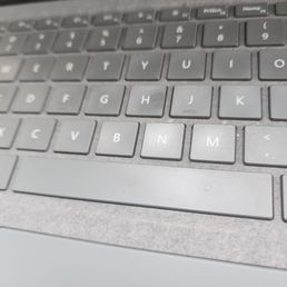 The image depicts a close-up view of a laptop keyboard with gray keys, including the letters B, N, M, and surrounding keys. The surface around the keyboard is also gray and appears to have some dust or smudges. The top part of the image shows part of the laptop's screen bezel. The photo is taken at an angle, focusing on the central keys.