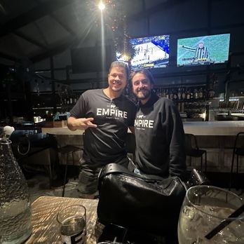 Empire - Two men wearing black Empire-branded shirts stand close together in a bar or restaurant with a dark, industrial-style interior. Behind them, a bar counter with bottles and two TV screens showing sports are visible. The table in front has glasses, a water pitcher, and a black bag. The setting appears lively with dim lighting and a festive atmosphere. - Burgers Near Me - Naperville, Illinois