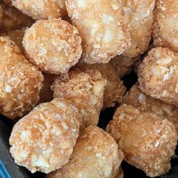 $2 tots - The image shows a close-up view of multiple fried chicken nuggets with a golden-brown, crispy breaded coating. They are placed in a black container, likely prepared for serving or eating. The nuggets are irregularly shaped and appear crunchy and well-cooked.