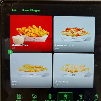 Shake Shack - A digital menu screen at Shake Shack in Toronto displays four fries options: Shaokao Fries priced at $5.99, Bacon Cheese Fries at $7.49, Fries at $4.49, and Cheese Fries at $5.99. The screen features colorful images of each dish, with the menu options arranged in a grid format. The top of the screen includes navigation options such as 'Exit' and 'Show Allergies,' and the bottom has icons for different food categories like burger, chicken, and shakes. - Hot Dogs Near Me - Toronto, Ontario