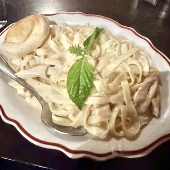 Tony C's - Fettuccini Alfredo with chicken - A serving of fettuccini Alfredo with chicken is presented on a decorative oval plate, topped with a sprig of basil and a basil leaf. A bread roll is placed on the side of the plate. The dish appears creamy and is set on a dark wooden table, with a metal spoon resting on the plate. The setting suggests a restaurant environment, possibly Italian cuisine. - Pizza Near Me - Austin, Texas