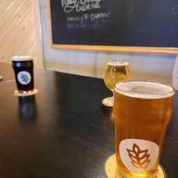 Three glasses of beer are arranged on a dark table inside a brewery, each on a round coaster with a leaf logo. The beers vary in color from light to dark. A chalkboard menu with handwritten text is visible in the background, indicating the brewery's name and offerings. The setting features wood-paneled walls, creating a casual, indoor atmosphere.