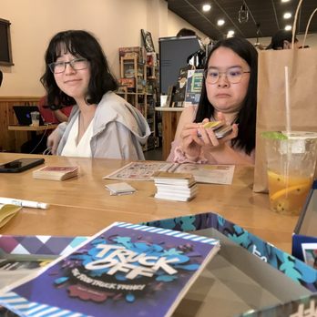 Squarrel Cafe - Love the vibes here! - Two young women with glasses sit at a table in a cafe, playing a card game with game cards and a game box visible. - Coffee & Tea Near Me - Brooklyn, New York