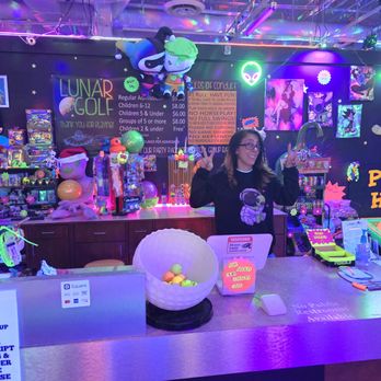 Lunar Golf - A woman stands behind a counter at Lunar Golf, an indoor mini-golf facility with vibrant neon lighting and space-themed decorations. The background features plush toys, colorful artwork, and a sign indicating a prize hole. The counter has various items, including a bowl of balls, a sign about loyalty, and a card reader. The environment is decorated with glowing alien and UFO motifs, creating a playful, extraterrestrial atmosphere. - Mini Golf Near Me - Albuquerque, New Mexico