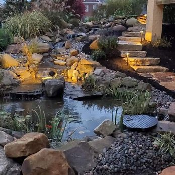 Rosenberger Scapes & Contracting - A natural stone walkway and steps with boulders, a waterfall, mulch, river rock, and lights. - General Contractors Near Me Rosenberger Scapes & Contracting - A natural stone walkway and steps with boulders, a waterfall, mulch, river rock, and lights. - General Contractors Near Me