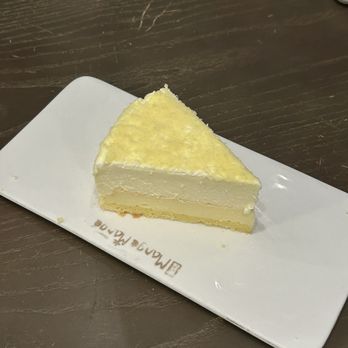 Mango Mango Dessert - Double Fromage Cheesecake (Slice) - A slice of cheesecake with a creamy filling and a light yellow topping is placed on a white rectangular plate on a dark wooden table. - Desserts Near Me - Edison, New Jersey