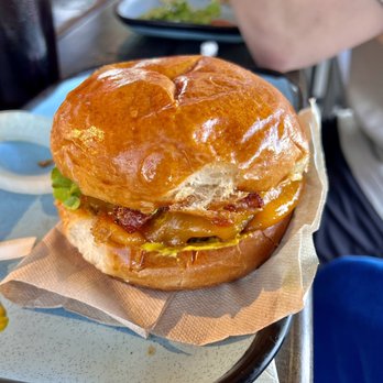 Twisted Root Burger - The Classic - A burger features a glossy, toasted bun with a beef patty topped with melted cheddar cheese, crispy bacon, lettuce, and pickles. The burger is placed on a paper-lined plate, with a small dish of sauce nearby. The background shows a blurred view of a dining setting with a person’s arm and a plate of salad. - Burgers Near Me - Abilene, Texas