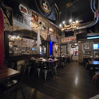 SafeHouse - One of the dining rooms - The image shows the interior of a themed restaurant with vintage decor, including brick walls adorned with flags, posters, and framed photographs. Wooden tables are set with napkins and utensils, and metal chairs surround them. The ceiling features a chandelier and decorative piping, with a large flag hanging prominently. The lighting is subdued, creating a cozy atmosphere. - Burgers Near Me - Milwaukee, Wisconsin