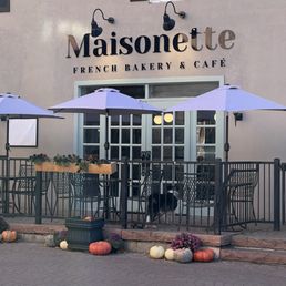 The image shows the exterior of Maisonette, a French bakery and café, with a sign above the entrance. The outdoor seating area includes tables, chairs, and umbrellas, enclosed by a black metal fence. Decorative pumpkins and flowers are placed along the front of the patio, and a dog is standing near the entrance. The building has a window with black shutters and a neutral-colored wall.