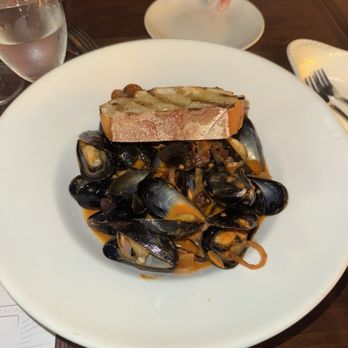 Blue Collar - Miss - A white plate holds a serving of cooked mussels in a sauce, garnished with caramelized onions. A slice of toasted bread rests on top of the mussels. The setting includes a glass of water, a fork, and a napkin on a wooden table, with a person’s hand and part of their arm visible in the background. - Comfort Food Near Me - Miami, Florida
