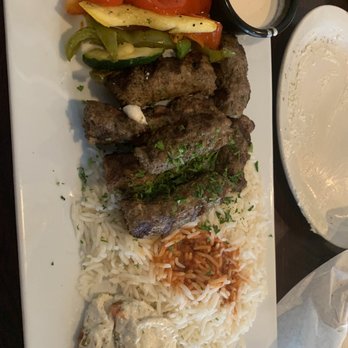 Aladdin Cafe - Lamb Kabob - The image shows a rectangular white plate with grilled meat garnished with herbs, white rice topped with tomato sauce, sautéed mixed vegetables including peppers and zucchini, and a side of creamy sauce with vegetables. The plate is placed on a dark wooden table, with a small black container of sauce and a white bowl or plate nearby. The setting suggests a Mediterranean or Middle Eastern cuisine restaurant, Aladdin Cafe in Lawrence, KS. - Greek Near Me - Lawrence, Kansas