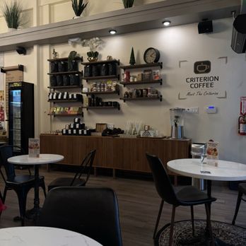 Criterion coffee - The photo depicts the interior of Criterion Coffee, with a wall-mounted shelving unit holding coffee bags, cans, and decorative items. There are tables with chairs arranged for seating, and a high shelf with potted plants above. The decor includes a clock, a sign with the Criterion Coffee logo, and lighting fixtures. The space has a modern, cozy atmosphere with wood flooring and a neutral color palette. - Coffee & Tea Near Me - Pasadena, California
