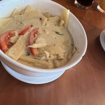 Thai Tip - Green Curry - A white ceramic bowl filled with green curry featuring chunks of chicken, sliced tomatoes, and vegetables in a creamy, greenish sauce. The bowl is on a wooden table, with other dishes and drinks partially visible around it, indicating a restaurant setting. - Thai Near Me - Albuquerque, New Mexico