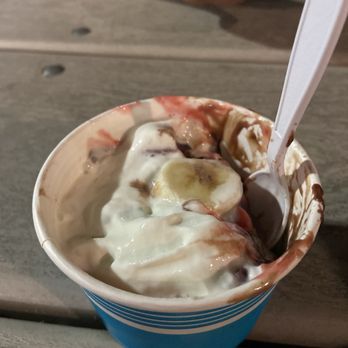Two Scoops - Sundae that I couldn't wait to eat! - A paper cup filled with ice cream topped with banana slices, chocolate syrup, and whipped cream is shown on a wooden table. A plastic spoon is inserted into the ice cream, and part of a person's hand is visible in the background. The setting appears to be outdoors or in a casual dining area. - Ice Cream & Frozen Yogurt Near Me - Osseo, Minnesota