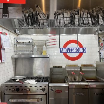 Above Ground - The image shows a commercial kitchen with stainless steel appliances including a stove, fryers, and shelving units. A sign with the word 'ABOVEGROUND' is mounted on a white tiled wall, which also displays a red and white logo. Above the kitchen area, there are wire baskets containing packaged items, and a red 'Open' sign is visible in the upper left corner. The setup suggests a food service environment, possibly within a food hall or restaurant. - Fish & Chips Near Me - Washington, DC, District of Columbia
