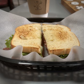 Blackjack’s - A toasted sandwich with visible lettuce and meat is placed on a black tray lined with parchment paper. The sandwich bread appears golden brown and toasted. In the background, there is a paper coffee cup with a logo, placed on a table with a wooden surface and some white coasters. The setting suggests a casual dining environment, possibly a coffee shop or café. - Coffee & Tea Near Me - Dublin, Texas