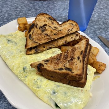 Bea's Cafe - an omelet with toast and potatoes - A breakfast plate includes a folded omelet with visible herbs, two slices of French toast with a marbled appearance, and a side of home fries. The meal is served on a white dish placed on a gray speckled countertop, with a blue water glass and a red bowl with condiments in the background. - Breakfast & Brunch Near Me - Conway, New Hampshire