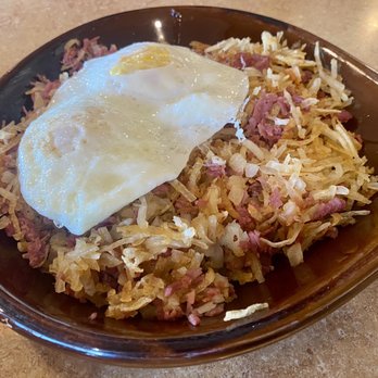 Weck's - Corn beef hash with two eggs over easy, requested hash made crisp! - The image shows a dish of hash topped with a fried egg, served in a brown, boat-shaped dish at Weck's in Santa Fe, NM. The hash contains shredded potatoes, ham, and possibly other ingredients, with the egg cooked sunny-side-up. The dish is placed on a beige countertop, and part of another dish is visible on the left side of the image. - Breakfast & Brunch Near Me - Santa Fe, New Mexico
