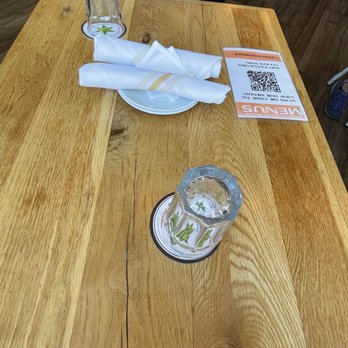 Uncommon Ground - Table - The image shows a wooden table in a restaurant setting, with two empty glasses placed on coasters, a stack of white napkins, and a menu with a QR code. A person wearing a blue shirt and black jacket is seated at the table, with part of their torso visible. The background includes a wooden floor and chairs, indicating an indoor dining area. - Breakfast & Brunch Near Me - Chicago, Illinois