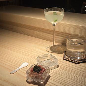 Ikigai - Toro tartare and martini - A cocktail glass with a clear beverage and a lime wedge inside is positioned on a wooden bar or table, alongside a glass of water on a coaster. In front of the drinks, there is a small square glass dish containing a dessert topped with berries and a tiny spoon nearby. The background features a dark, wooden interior with subtle lighting, suggesting an upscale bar or restaurant environment. - Japanese Near Me - Brooklyn, New York