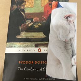 Bookmark of sleeping Lyla! - The image shows a hardcover book titled "The Gambler and Other" by Fyodor Dostoevsky, published by Penguin Classics. The book is placed on a flat surface, and a white cat with closed eyes is sleeping on top of it, partially covering the cover. The cat appears relaxed and is resting comfortably, with its head and paws visible. The background of the book cover features an illustration of two men sitting at a table, engaged in a card game or conversation.