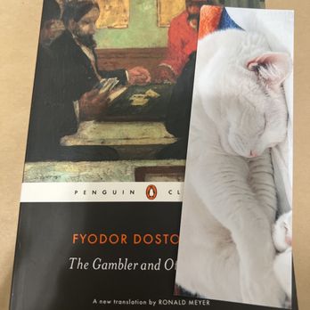 Sleepy Cat Books - Bookmark of sleeping Lyla! - The image shows a hardcover book titled "The Gambler and Other" by Fyodor Dostoevsky, published by Penguin Classics. The book is placed on a flat surface, and a white cat with closed eyes is sleeping on top of it, partially covering the cover. The cat appears relaxed and is resting comfortably, with its head and paws visible. The background of the book cover features an illustration of two men sitting at a table, engaged in a card game or conversation. - Bookstores Near Me - Berkeley, California