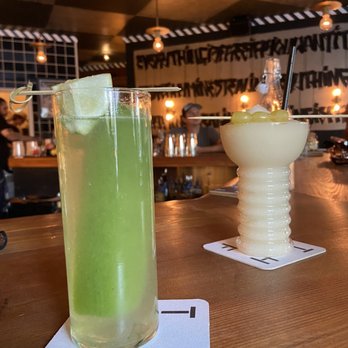 Thief - The Supersonic and Friesling - The image shows two cocktails on a wooden bar counter inside a bar with graffiti-style wall art and warm ambient lighting. The green cocktail is in a tall glass with ice and a garnish, while the white cocktail is in a textured, ribbed glass with a straw and fruit garnish. The background features bar staff and patrons, with a ceiling decorated with checkered patterns and hanging lights. - Bars Near Me - Brooklyn, New York