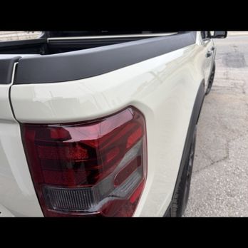 Carfection Pro - After, absolutely flawless! - A photograph captures the rear left side of a white pickup truck parked on a paved surface. The image prominently features the taillight assembly, which includes red and clear sections, and part of the truck bed with a black trim. The background shows a section of the pavement and a clear sky. The photo emphasizes the vehicle's exterior details and condition. - Body Shops Near Me - Oxnard, California