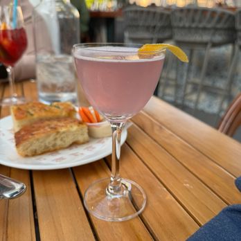 Celeste - Celeste 75 - A pink cocktail in a coupe glass with a lemon twist garnish is placed on a wooden table at an outdoor dining area, with food and drinks visible in the background. - Desserts Near Me - Narragansett, Rhode Island