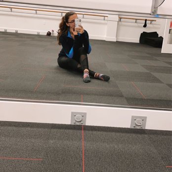 Pure Barre - Pre-Class Photo at the Pure Barre Appleton Studio - A woman with glasses and long hair, dressed in athletic clothing, sits on the floor of a dance studio, holding her phone to take a mirror selfie. The studio features a large wall mirror, ballet barres mounted on the wall, and a carpeted floor with red grid lines. The background includes a white wall, some black equipment, and a wooden handrail above the mirror. The woman is wearing colorful socks and appears relaxed. - Barre Classes Near Me - Appleton, Wisconsin