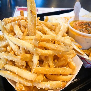 Nosh Shop - Fries - A tray of French fries topped with grated cheese, accompanied by a small container of dipping sauce, on a black tray. - Food Trucks Near Me - Eugene, Oregon