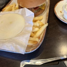 Burger with bacon and slice of raw onion - A burger with bacon and a slice of raw onion is placed on a tray alongside French fries. The tray also contains a cup of creamy dipping sauce or cheese sauce. The setting appears to be a casual dining restaurant with a dark wooden table. The burger has a toasted bun, and the fries are golden and slightly crispy. There is a small plate with a plastic cup and a spoon visible in the background.