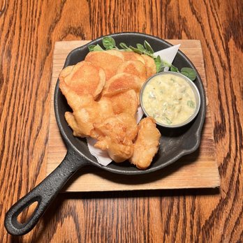 Michael's - Fish & Chips, Sauce Gribiche - A serving of fried fish and potato chips is presented in a small black skillet placed on a wooden table. The dish includes crispy battered fish pieces, golden potato chips, and a small cup of tartar sauce with chopped herbs. Green garnish is also included for presentation. The skillet is set on a wooden board, highlighting a casual dining presentation suitable for seafood or fish and chips offerings. - Seafood Near Me - St. Augustine, Florida
