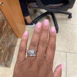 Pretty in Pink - A hand with pink manicured nails displays a diamond ring, with a nail salon setting in the background, including a person seated at a desk and a chair.