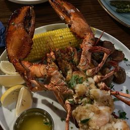 Stuffed Lobster - A seafood platter includes a split lobster tail, a whole lobster claw, grilled shrimp, a piece of corn on the cob, lemon wedges, and a small container of melted butter or oil, garnished with herbs. The dish is served on a white oval plate, with additional food items visible in the background, on a dark wooden table.