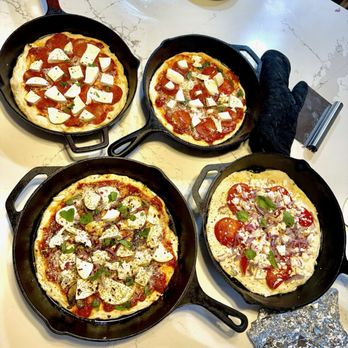 The Italian Diva - Four mini pizzas with different toppings are placed in cast iron skillets on a white marble countertop. The pizzas include ingredients such as cheese, pepperoni, vegetables, and fresh basil. A black oven mitt, a foil-wrapped item, and a pizza cutter are also visible on the counter. The setting suggests a casual pizza-making or tasting session, possibly in a kitchen or culinary class environment. - Cooking Classes Near Me - Wareham, Massachusetts