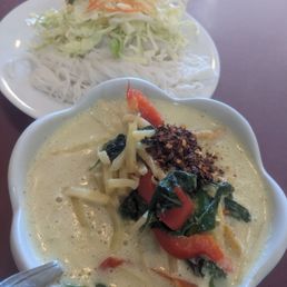 C1 Green Curry - A bowl of Thai green curry with vegetables and chili flakes is in the foreground, with a plate of noodles and shredded vegetables in the background.