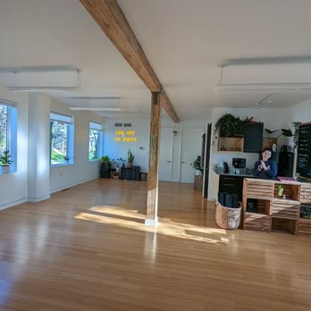 Collective Flow - It's a small modern, clean, organized studio to Collectively gather and "practice" Yoga! - A spacious, well-lit interior with wooden flooring, large windows, and a reception area with a person standing behind a counter, decorated with plants. - Yoga Near Me - Milwaukee, Wisconsin