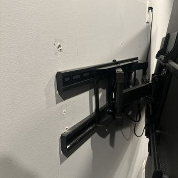 FixTman - Washington - Where technician had to take the bracket out of the wall and mount it lower - The image shows a black TV wall mount attached to a light-colored wall, with visible holes and damage around the mounting area. A white panel is partially visible on the wall, and a power cord hangs from the wall near the mount. The wall appears to have been drilled or altered, indicating ongoing or incomplete installation. - Furniture Assembly Near Me