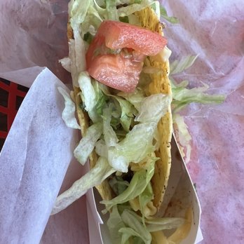 Toucan Taco - A taco with a crispy shell contains shredded lettuce, slices of tomato, and shredded cheese. It is served on a paper-lined tray, with some lettuce spilling out. The background includes a pinkish surface, and the taco appears to be freshly prepared, showcasing typical ingredients of a Mexican-style taco. - Mexican Near Me - Laurel, Maryland