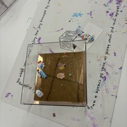 The image shows a small, square, reflective surface with a gold-colored finish, placed on a white table covered with colorful paint splatters. Several small unicorn and bear figurines are arranged on the reflective surface. Underneath, there is a piece of paper with handwritten text and a 2D DIY Studio logo. The background includes a transparent container with art supplies, indicating an arts and crafts setting.