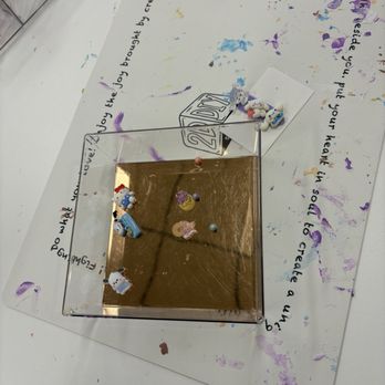 2D DIY Studio - The image shows a small, square, reflective surface with a gold-colored finish, placed on a white table covered with colorful paint splatters. Several small unicorn and bear figurines are arranged on the reflective surface. Underneath, there is a piece of paper with handwritten text and a 2D DIY Studio logo. The background includes a transparent container with art supplies, indicating an arts and crafts setting. - Art Classes Near Me - Troy, Michigan