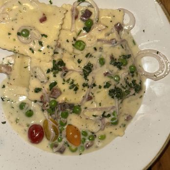 Basso - A plate of ravioli pasta is presented with a creamy white sauce, topped with green peas, cherry tomatoes, chopped herbs, and thinly sliced onions. The dish is served on a white plate with a subtle speckled pattern, and the background shows a wooden surface. - Bars Near Me - Saint Louis, Missouri