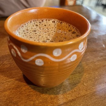 Delhicacy - Masala Chai - A close-up image of a clay cup filled with masala chai, showing foam on the surface. The cup has a decorative white pattern and is placed on a wooden table. The background suggests an indoor restaurant environment with blurred elements and warm lighting. - Indian Near Me - Cedar Rapids, Iowa