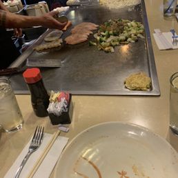 A teppanyaki-style restaurant scene with a chef preparing food on a flat-top grill, including rice, vegetables, and meat, with dining utensils and glasses on the table.