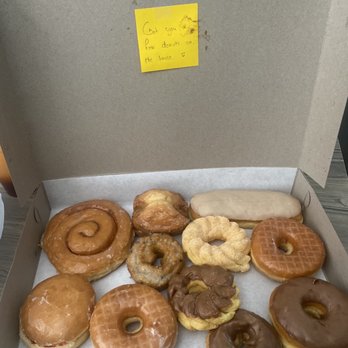 BC’s Donuts - A cardboard box holds an assortment of donuts, including glazed, chocolate-covered, and ring-shaped varieties. A yellow sticky note with handwritten text is attached to the inside of the box lid. The donuts are arranged on a sheet of parchment paper, and the box appears to be placed on a flat surface. - Donuts Near Me - Pasadena, California