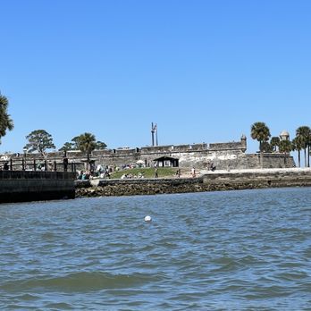 Bluewater Adventure - A historic stone fort with palm trees and visitors is seen across a waterway under a clear blue sky in St. Augustine, FL. - Boat Tours Near Me - St. Augustine, Florida